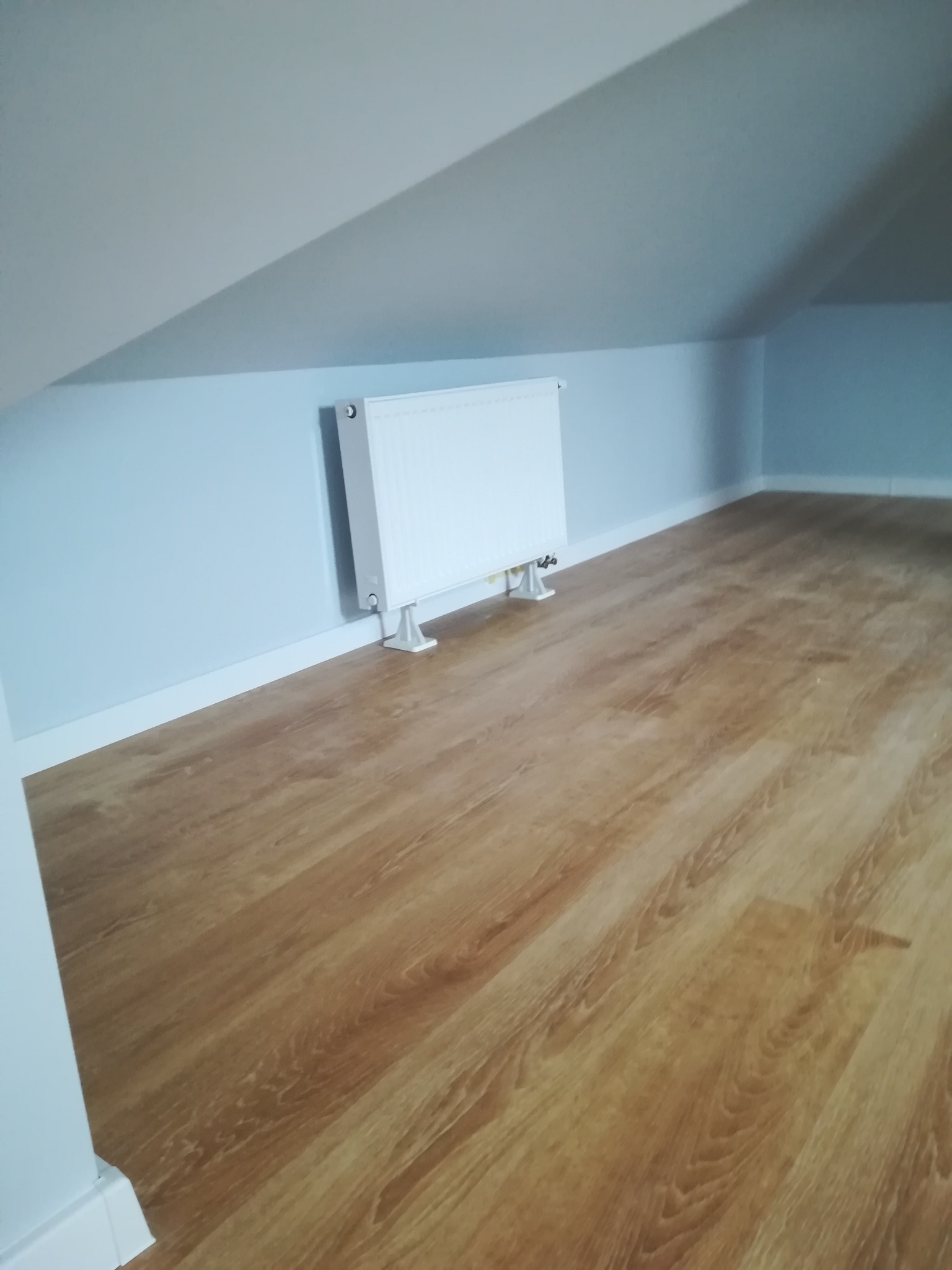 Poddasze po remoncie: jasne ściany, drewnopodobna podłoga i biały grzejnik.