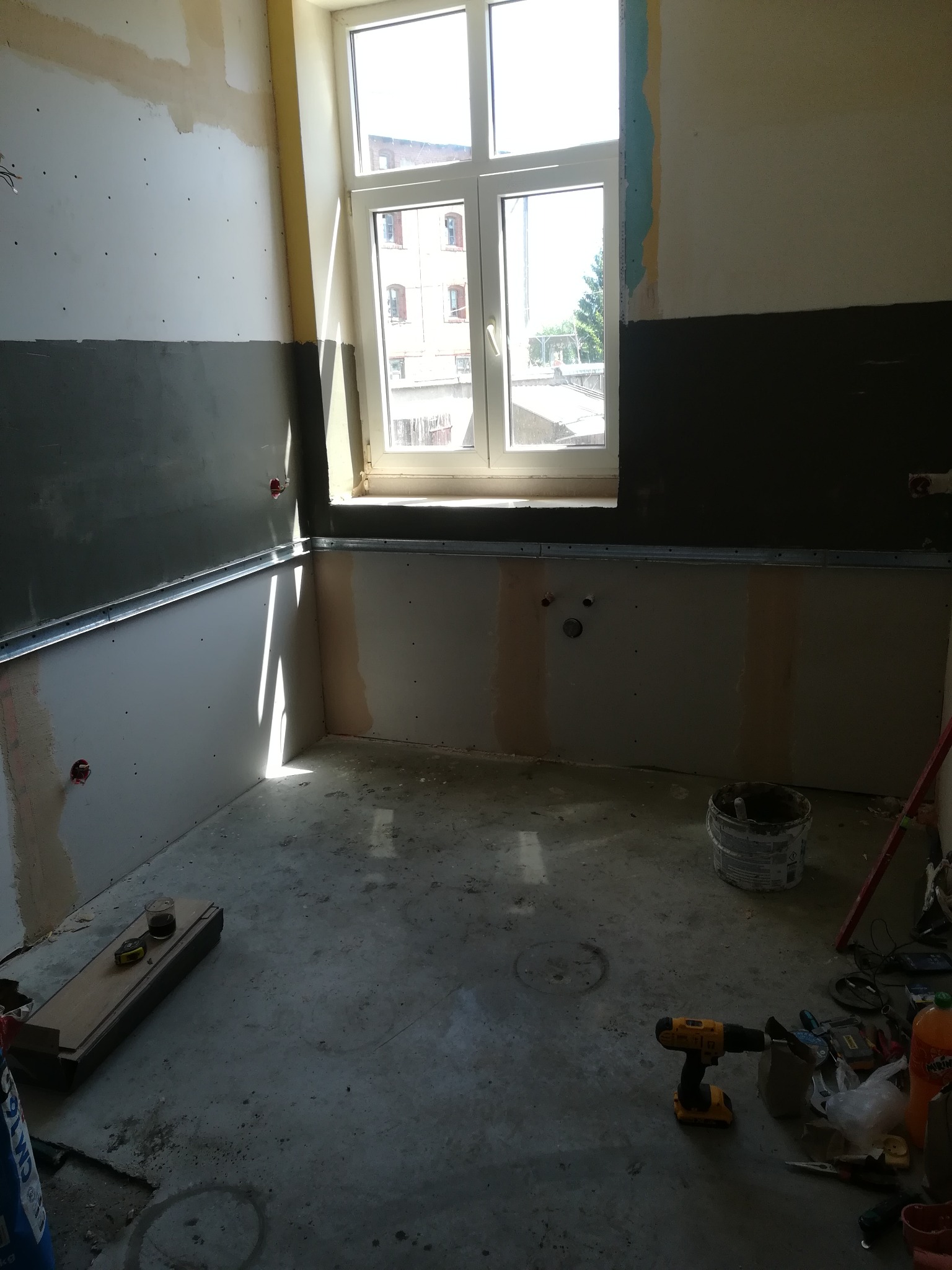Wnętrze w trakcie prac: ściany przygotowane pod montaż płyt, widoczne profile metalowe, częściowo pomalowane dolne fragmenty ścian na szaro, okno z widokiem na budynek mieszkalny, narzędzia...