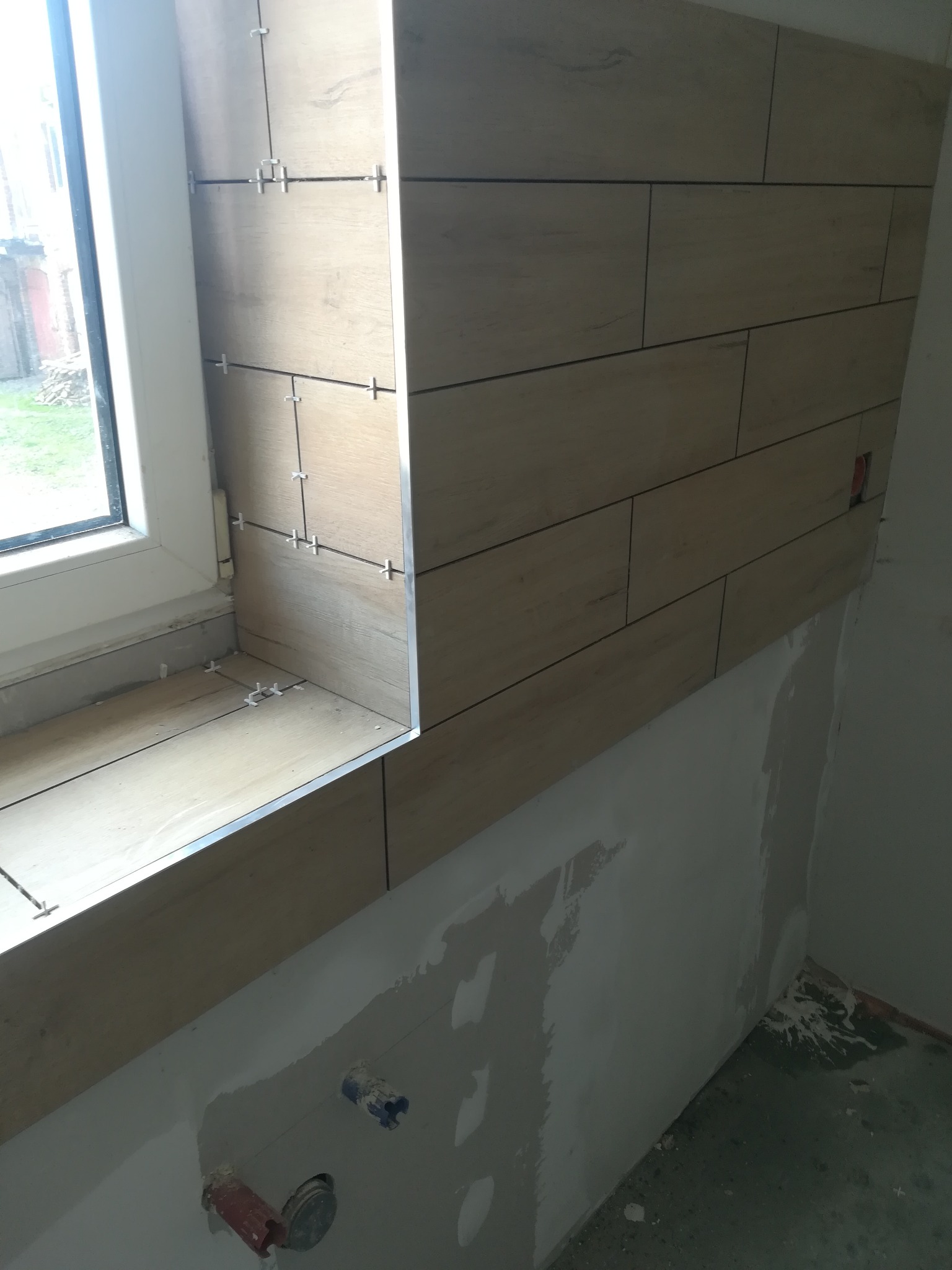 Układanie płytek imitujących drewno na ścianie i wnęce okiennej, widoczne krzyżyki dystansowe i fragment surowej ściany z instalacjami.