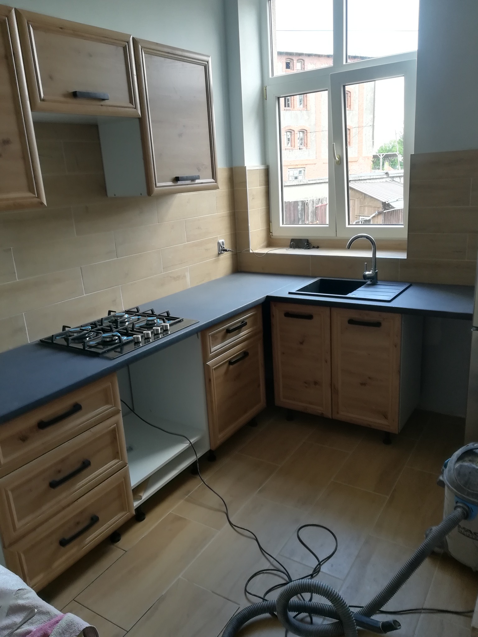 Jasna kuchnia z drewnianymi szafkami i czarnym blatem, widoczna płyta gazowa i zlew, płytki na ścianie, w tle okno z widokiem na budynek, na podłodze leży odkurzacz.