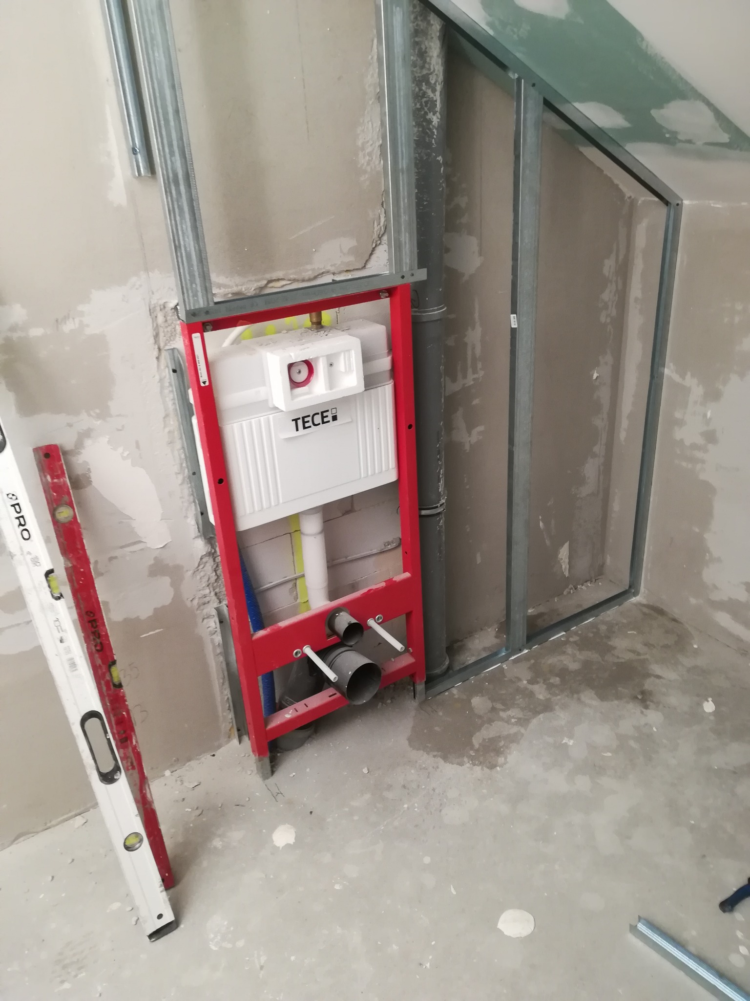 Stelaż podtynkowy WC marki TECE zamontowany na ścianie z profilami, obok poziomica PRO i fragment konstrukcji z płyty karton gips na poddaszu.