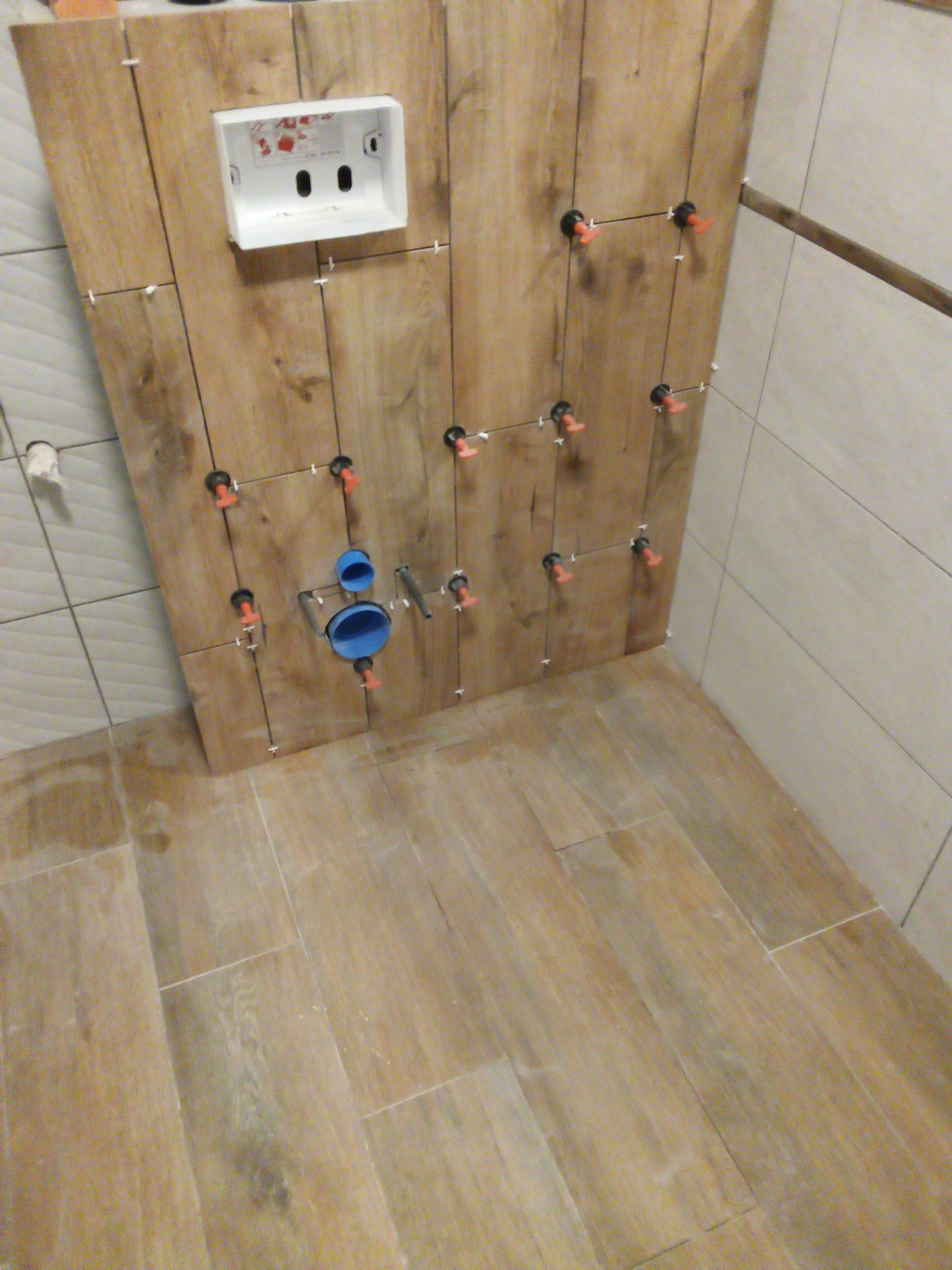 Montaż płytek imitujących drewno na ścianie, widoczna biała podtynkowa skrzynka WC i niebieskie rury kanalizacyjne, użyte systemy poziomowania płytek.