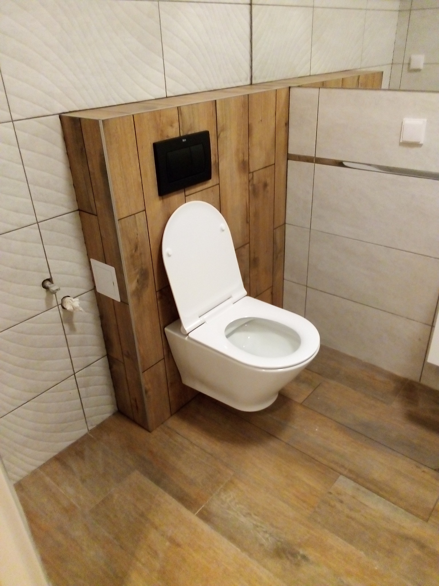 Nowoczesna toaleta podwieszana z drewnianą obudową stelaża i przyciskiem spłukującym, płytki imitujące drewno na podłodze i ścianie, widoczne przyłącze wody.