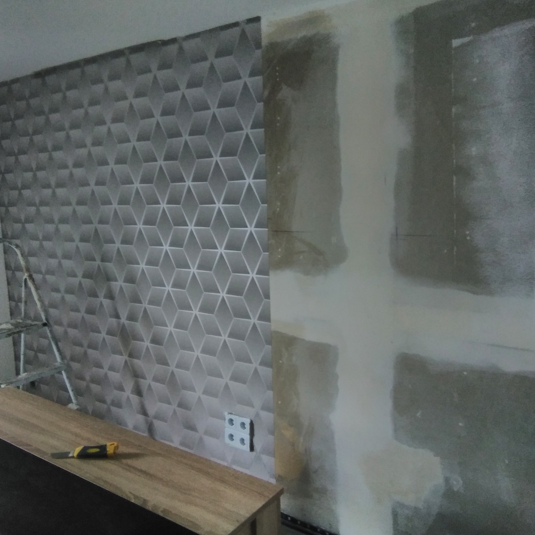 Fragment ściany: połowa pokryta tapetą w szaro-białe geometryczne wzory, druga połowa w trakcie szpachlowania, widoczna drabina i nóż do tapet.