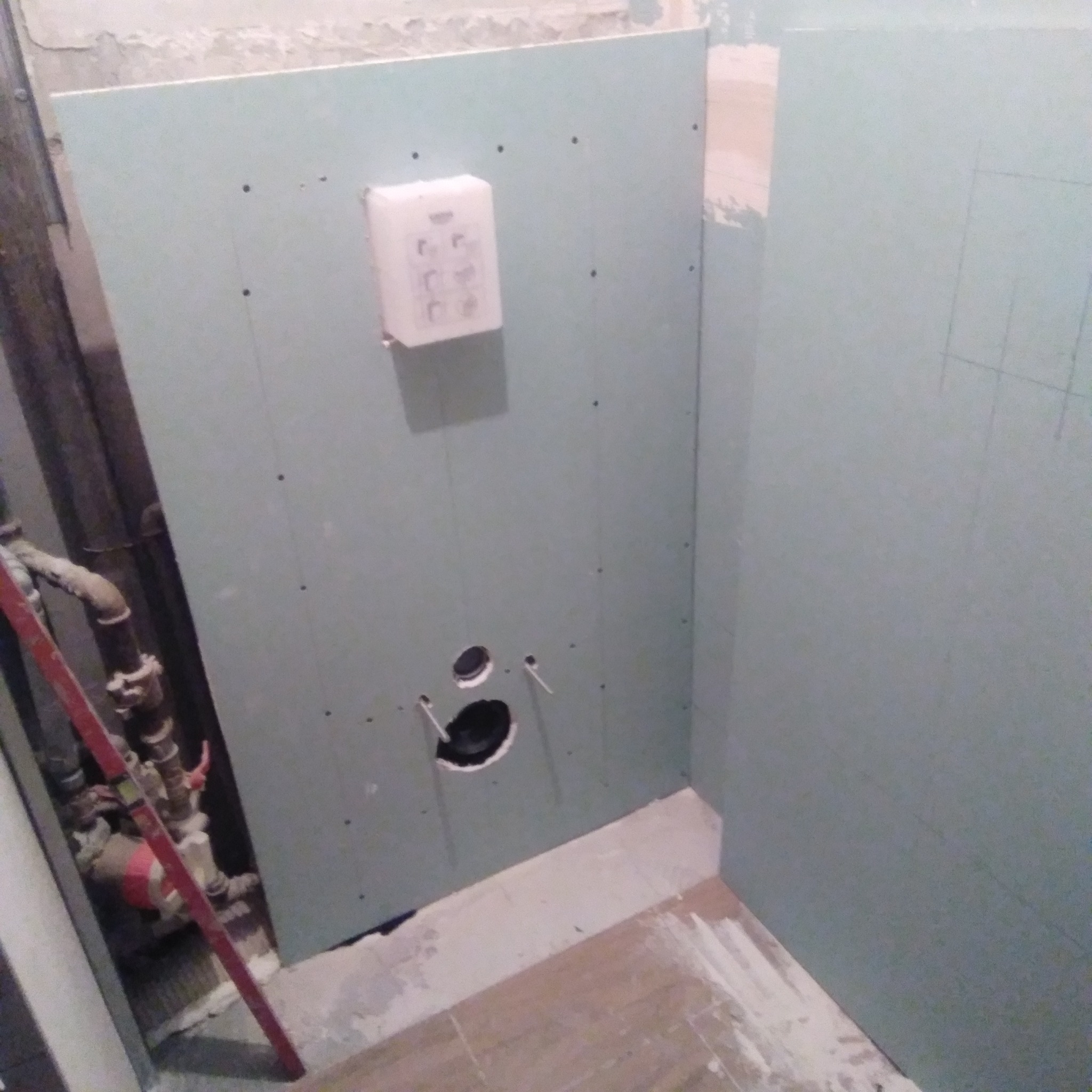 Montaż płyty karton gips w narożniku łazienki, widoczne otwory na instalacje, ślady ołówka na płycie, oraz instalacja wodna z zaworami i poziomicą oparta o ścianę.