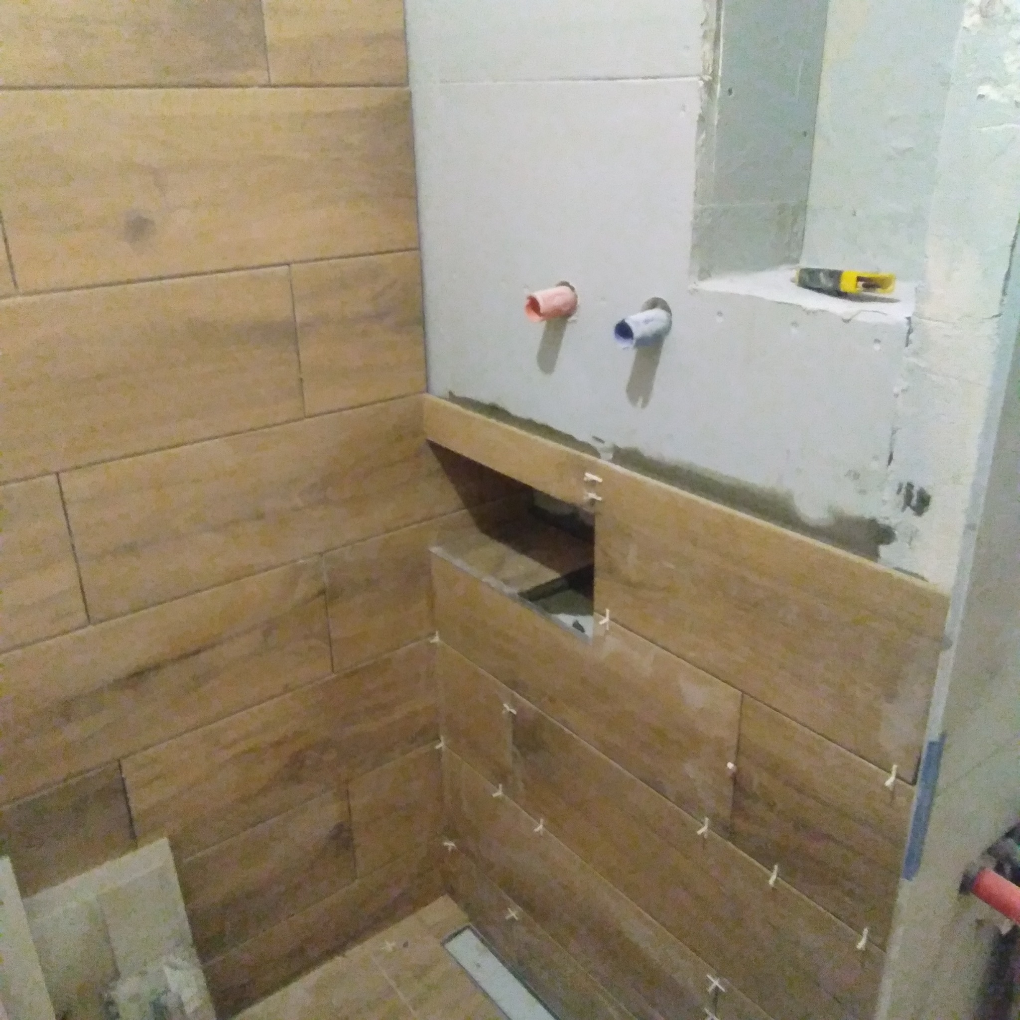 Montaż płytek imitujących drewno w łazience, widoczne krzyżyki dystansowe, przygotowane instalacje wodne i fragment narzędzia.