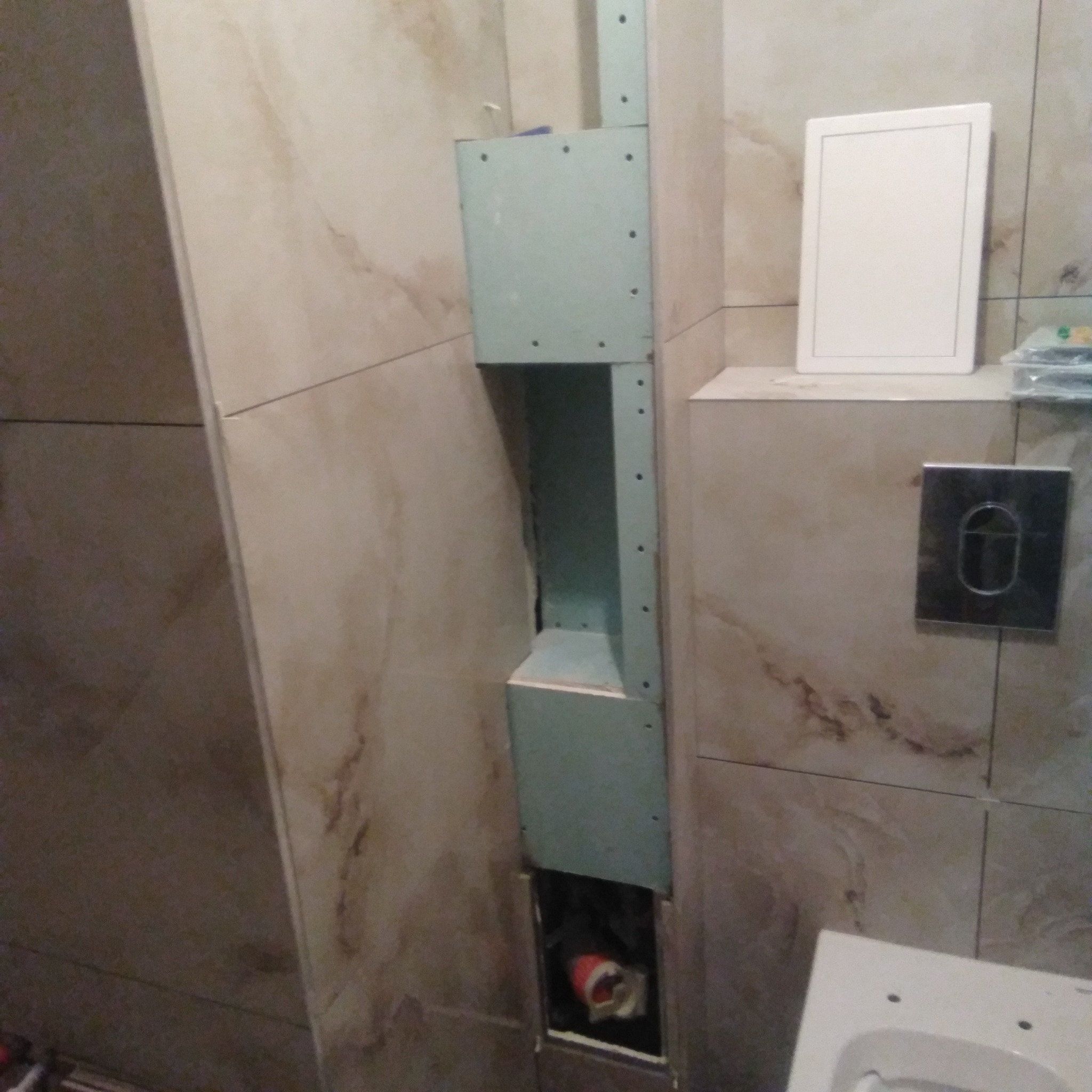 Konstrukcja z płyty karton gips zamontowana na ścianie z beżowych płytek, obudowująca instalację w łazience, widoczny fragment toalety i przycisk spłuczki.