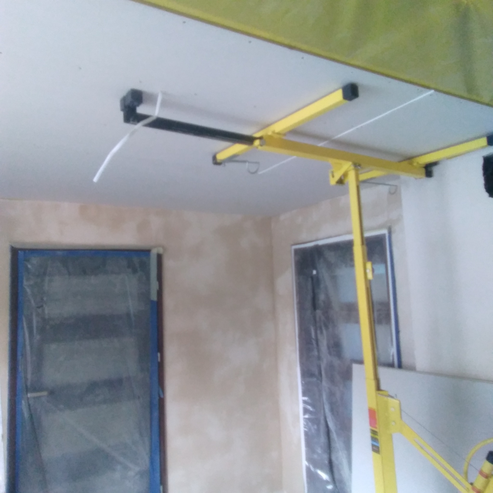 Montaż płyty kartonowo-gipsowej na suficie za pomocą żółtego podnośnika; widoczne zabezpieczone folią okna i ściana po wstępnym gipsowaniu.