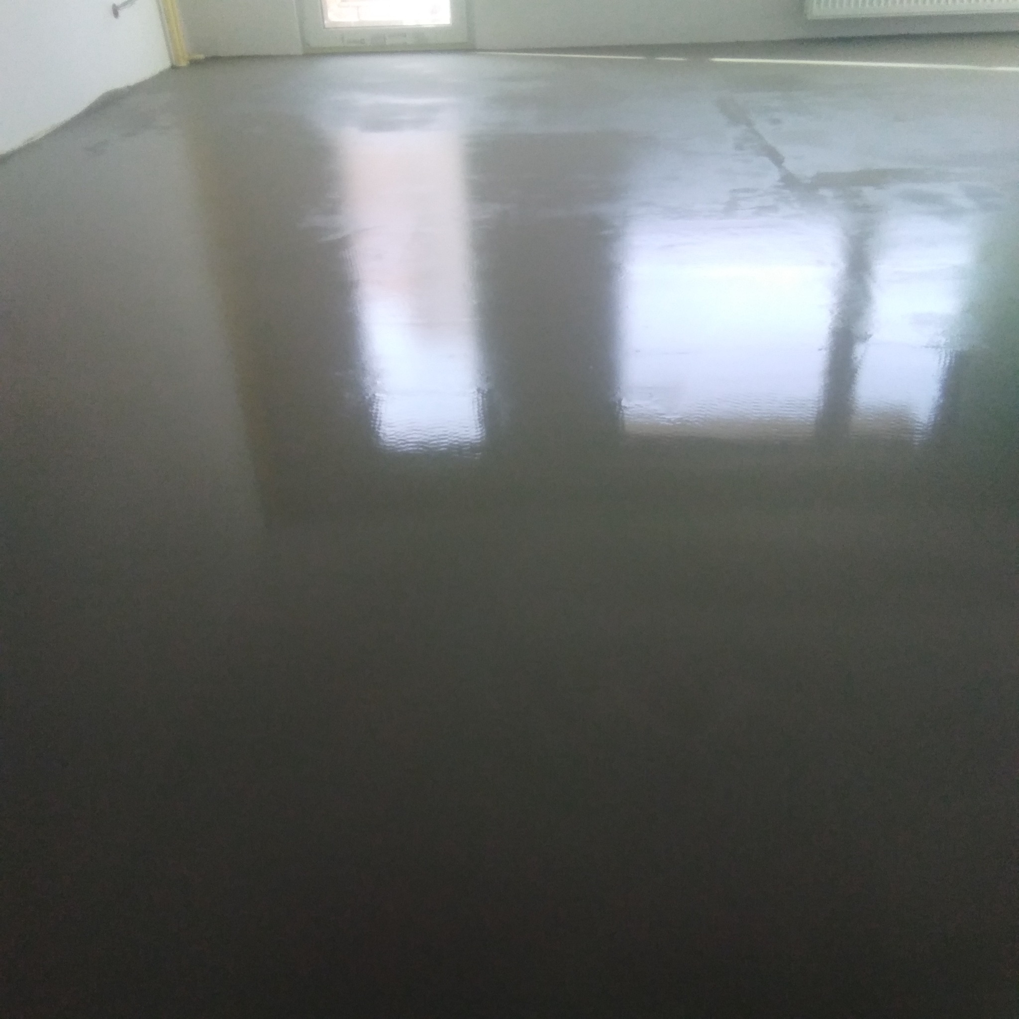 Wylany, mokry, samopoziomujący szary beton na podłodze w pokoju podczas prac wykończeniowych, z odbiciami okien i kaloryfera.
