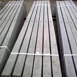 Betoniarstwo Agnieszka Kaczorowska - Palety szarych, betonowych słupków ogrodzeniowych, spiętych białym paskiem, ułożone równolegle na ziemi.