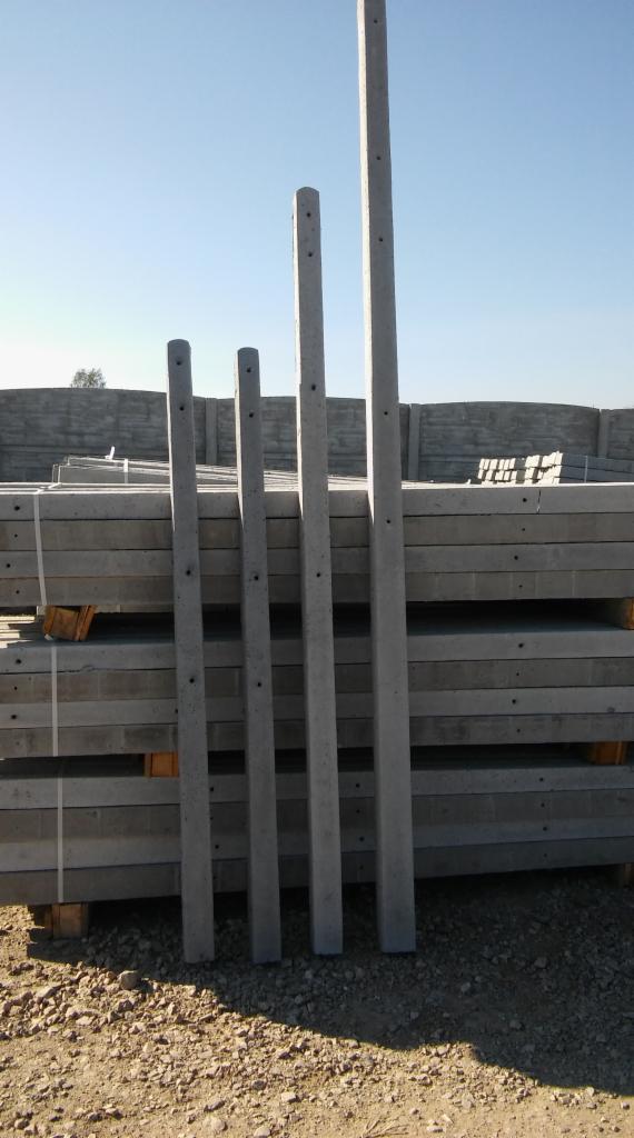 Betonowe słupki ogrodzeniowe ustawione pionowo przed stosem płyt betonowych na palecie, widoczne otwory montażowe, szare tło nieba.