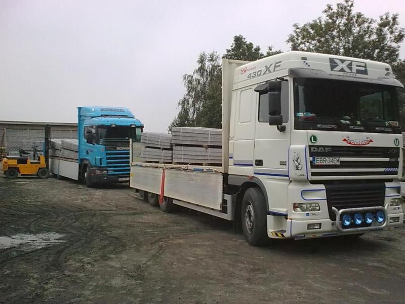 Dwa ciężarowe samochody, DAF i Scania, załadowane szarymi betonowymi płytami, z żółtym wózkiem widłowym w tle, na placu budowy.