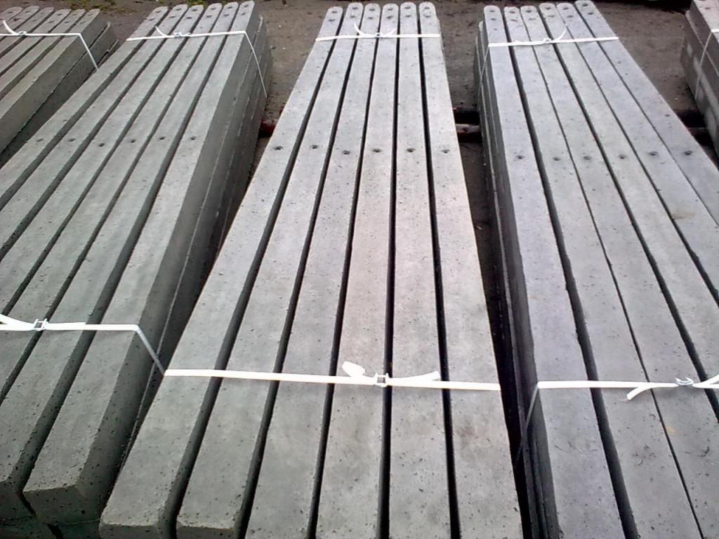 Palety szarych, betonowych słupków ogrodzeniowych, spiętych białym paskiem, ułożone równolegle na ziemi.