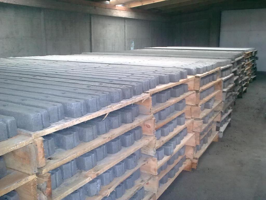 Szare betonowe słupki ogrodzeniowe ułożone na drewnianych paletach w magazynie, widoczne otwory montażowe w słupkach.