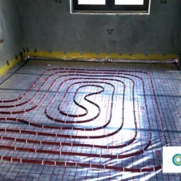 Widok na instalację ogrzewania podłogowego z czerwonymi rurkami ułożonymi w pętle na siatce, przygotowaną do zalania wylewką, w surowym wnętrzu z oknem i żółtą taśmą wzdłuż ścian.