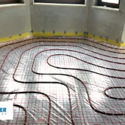 Instalacja ogrzewania podłogowego w trakcie montażu: rury grzewcze ułożone na folii z rastrem, mocowane taśmą do ścian, widoczne okna i surowy tynk.