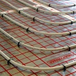 Białe rury ogrzewania podłogowego zamocowane czarnymi uchwytami na srebrnej folii z czerwoną kratą, widok z góry.