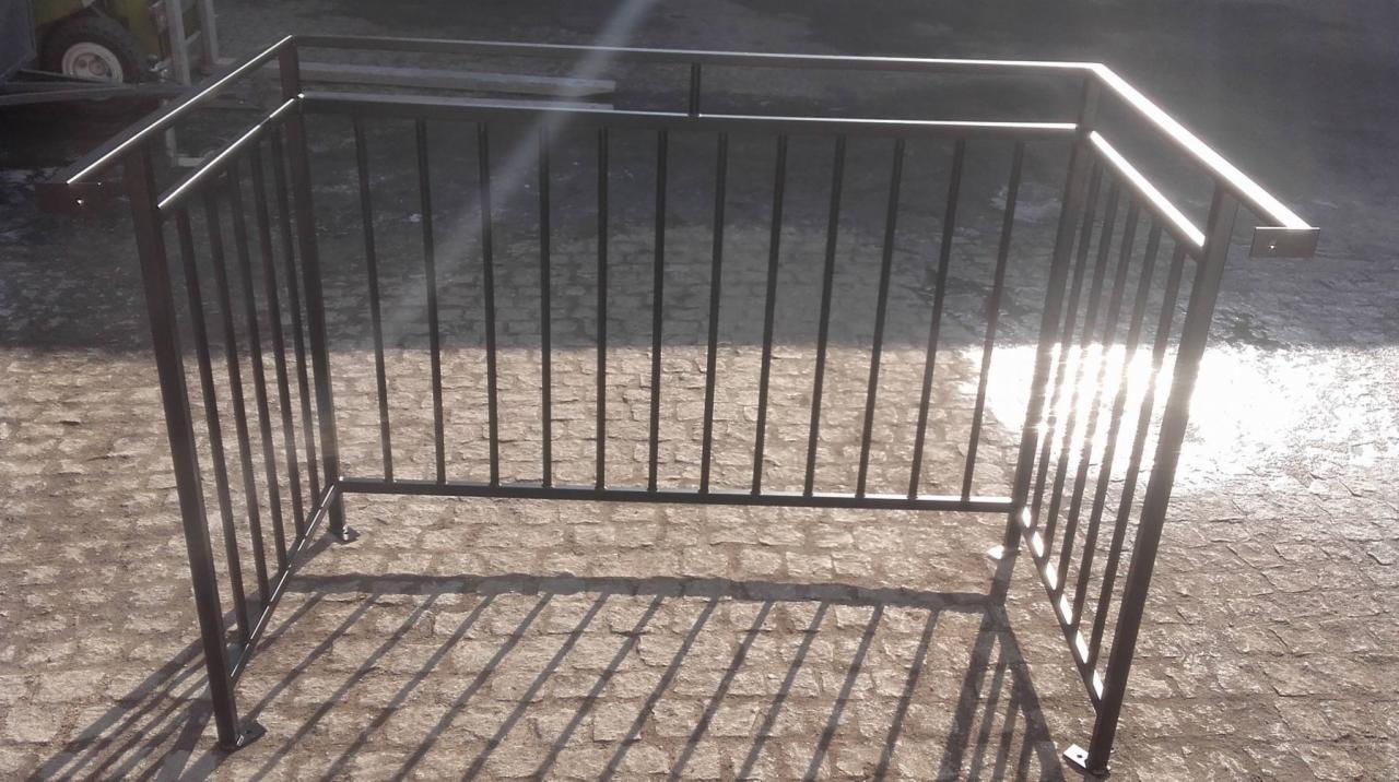 Metalowa konstrukcja balustrady w kolorze grafitowym, spawana z profili zamkniętych, ustawiona na brukowanej powierzchni, z widocznymi spawami w narożach i odbiciami słońca na powierzchni.