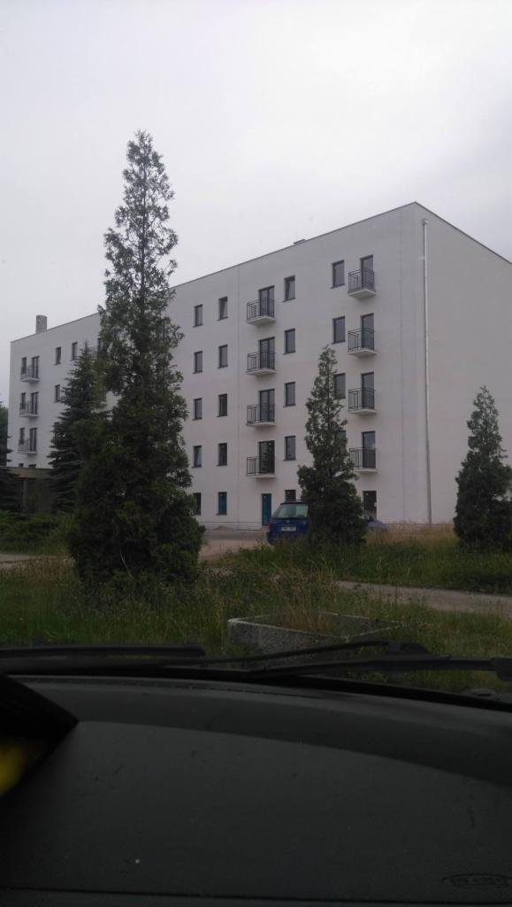 Nowo wybudowany, otynkowany na biało blok mieszkalny z balkonami, widziany zza przedniej szyby samochodu, z drzewami iglastymi na pierwszym planie i zaparkowanym niebieskim samochodem w tle.