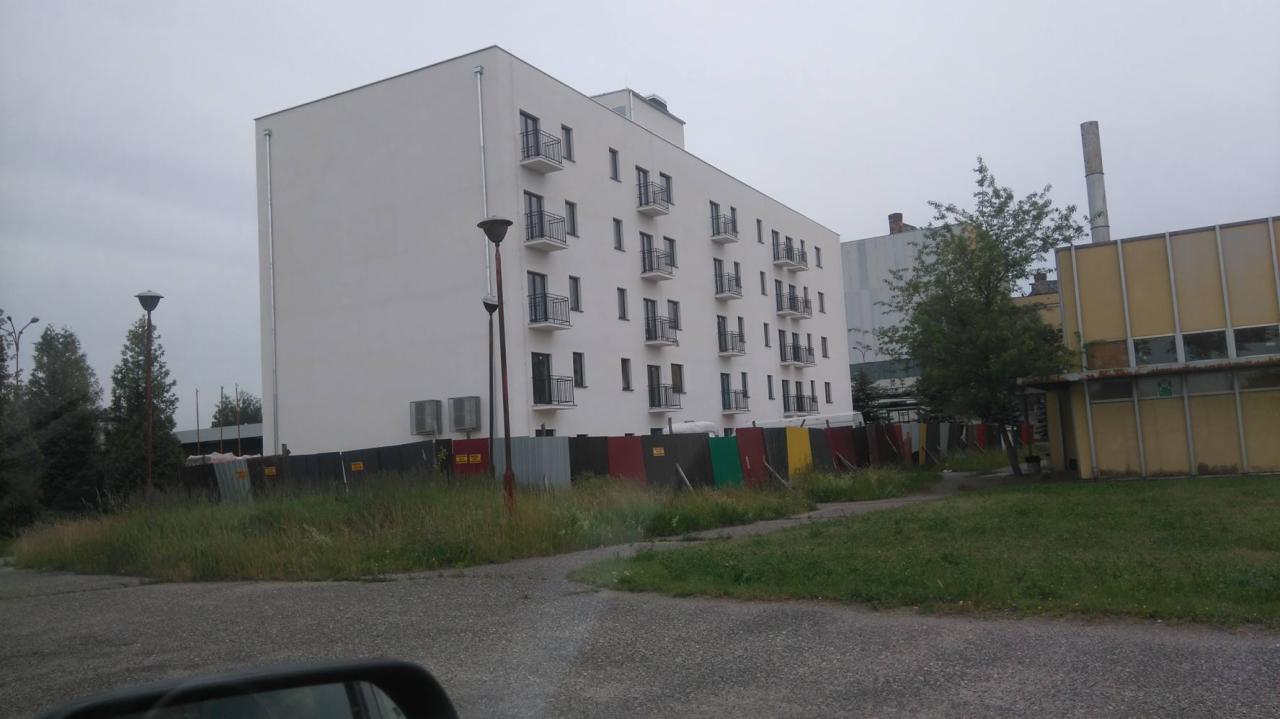 Nowoczesny, biały budynek wielorodzinny z balkonami, widoczny zza tymczasowego ogrodzenia budowlanego z kolorowych blach, w tle komin przemysłowy i budynek o żółtej elewacji.