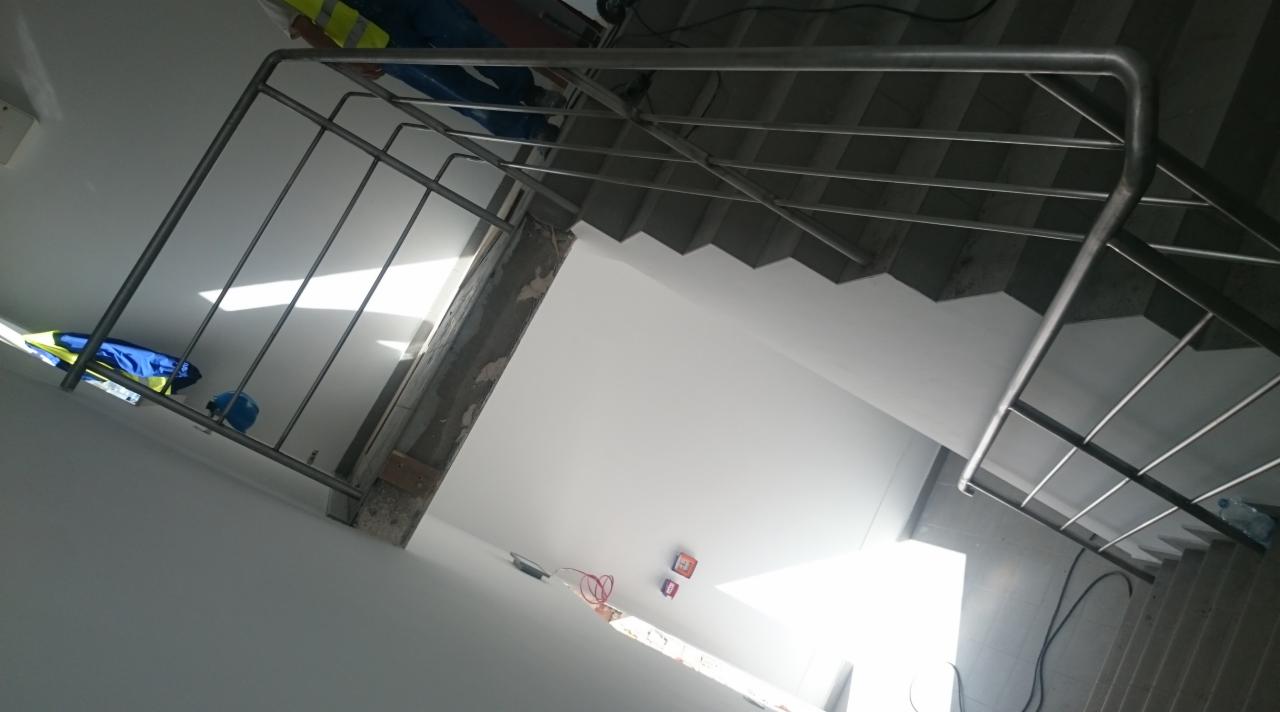 Metalowa balustrada schodowa z wypełnieniem prętowym, widoczna z dołu, z elementami odzieży roboczej i kaskiem w tle.