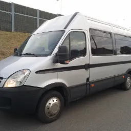 Srebrny bus pasażerski Iveco z przyciemnianymi szybami, zaparkowany na asfalcie, z widocznym fragmentem zielonego zbocza i ekranu akustycznego w tle.