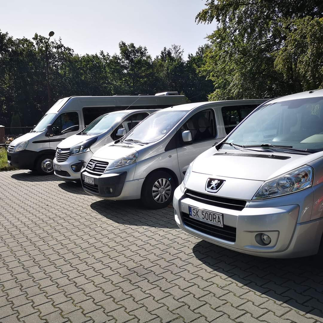 Flota srebrnych busów i vanów zaparkowana na brukowanym parkingu, z widocznymi numerami rejestracyjnymi i drzewami w tle.