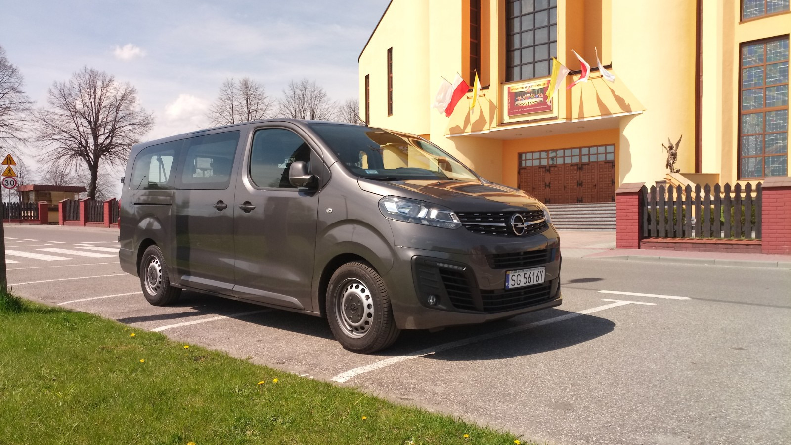 Szary Opel Zafira Life zaparkowany na parkingu w Zabrzu, na tle żółtego budynku z polskimi flagami.