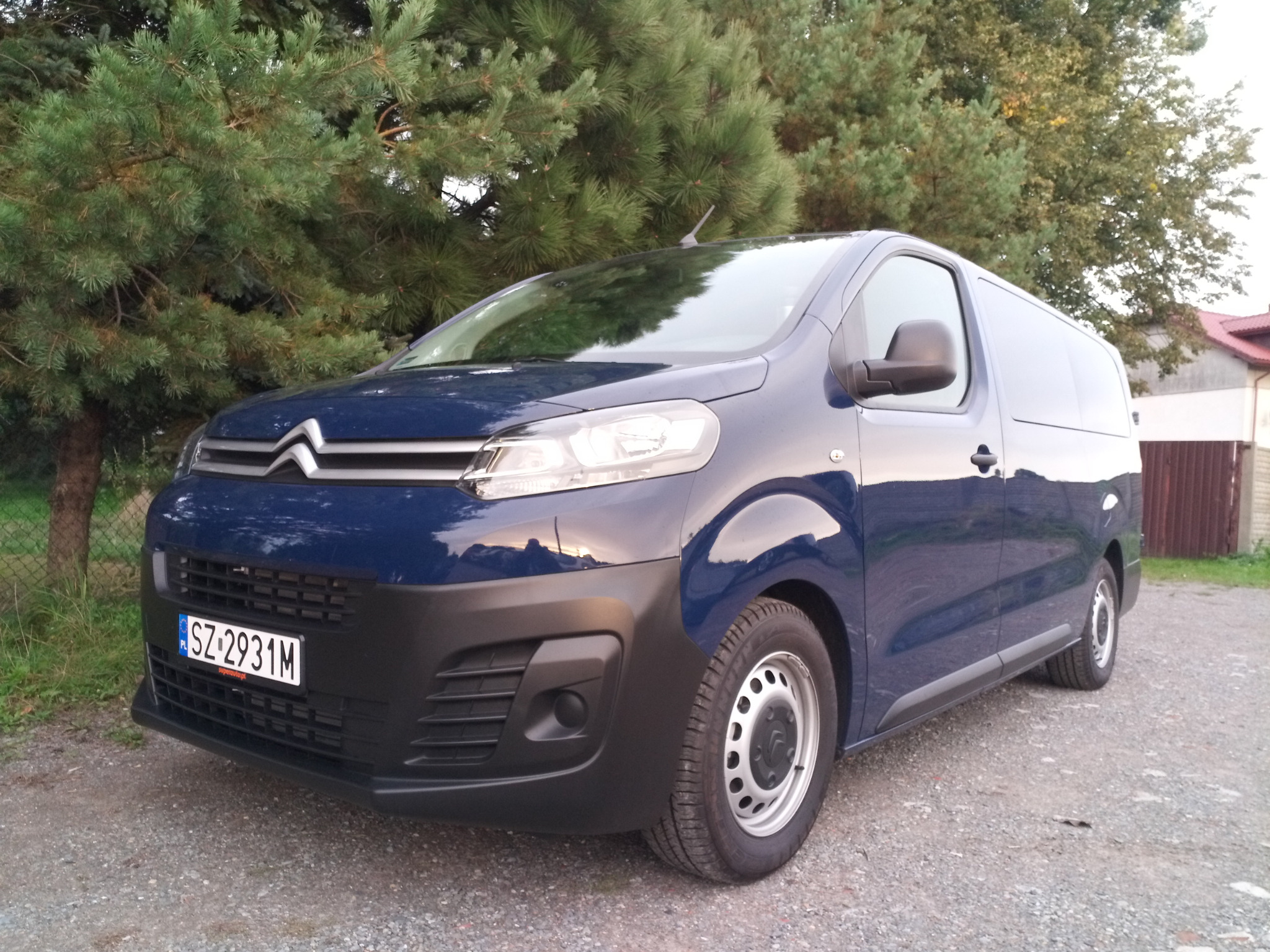 Niebieski samochód dostawczy Citroen z polską tablicą rejestracyjną, zaparkowany na żwirowej powierzchni, na tle zielonych drzew.