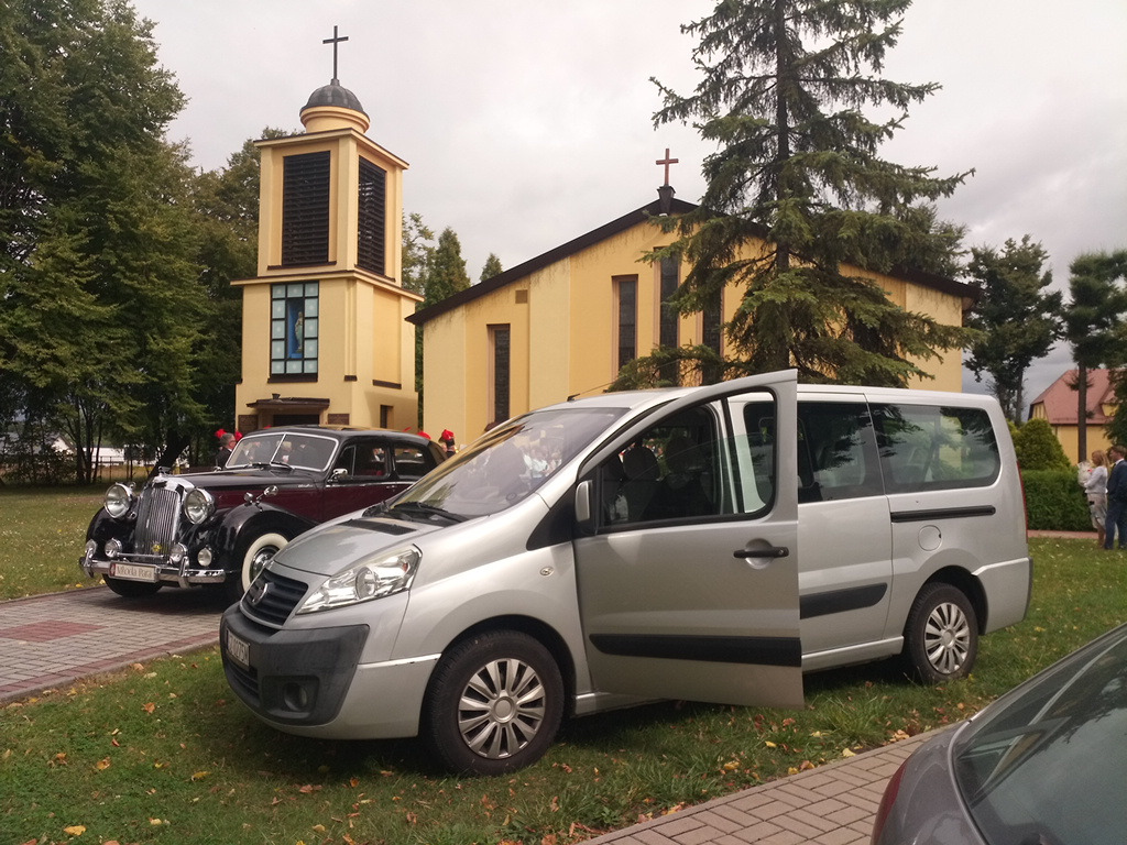 Srebrny van z otwartymi drzwiami na tle kościoła i klasycznego, czarnego samochodu z chromowanymi elementami, prawdopodobnie podczas ceremonii ślubnej.