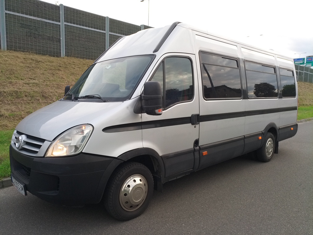 Srebrny bus pasażerski Iveco z przyciemnianymi szybami, zaparkowany na asfalcie, z widocznym fragmentem zielonego zbocza i ekranu akustycznego w tle.