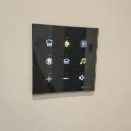 Szklany panel dotykowy inteligentnego domu z ikonami sterowania oświetleniem, temperaturą, muzyką i innymi funkcjami, zamontowany na jasnej ścianie.