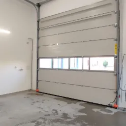 Zamknięta, segmentowa brama garażowa w jasnym kolorze z poziomymi panelami i rzędem okien, widoczna od wewnątrz garażu. Na posadzce widoczne ślady obuwia. Brama wyposażona w napęd elektryczny.