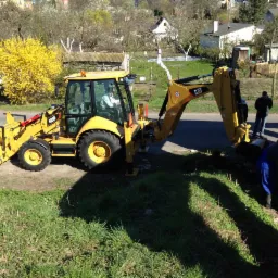 Żółta koparko-ładowarka CAT 428F podczas prac ziemnych przy budowie ściany z czerwonej cegły, w tle zabudowa jednorodzinna i kwitnący krzew forsycji.