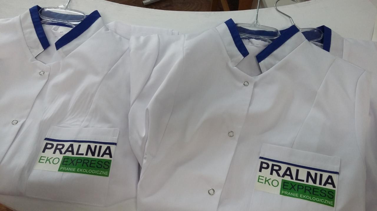Białe, nowo uszyte fartuchy robocze z granatowymi wykończeniami i logo pralni Eko Express, wiszące na wieszakach.