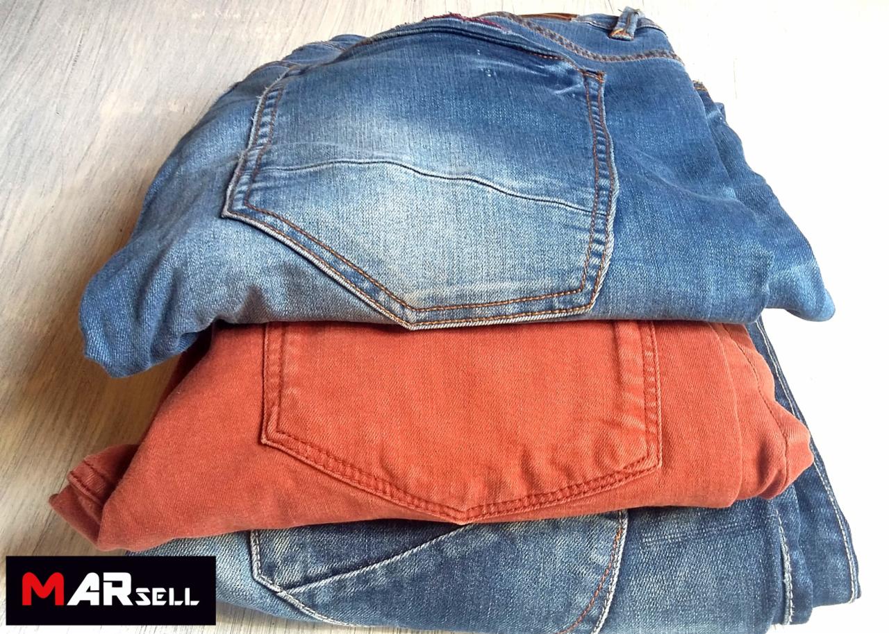 Stos trzech par złożonych jeansów: dwie pary niebieskich i jedna pomarańczowa, widoczne tylne kieszenie, logo firmy w lewym dolnym rogu.