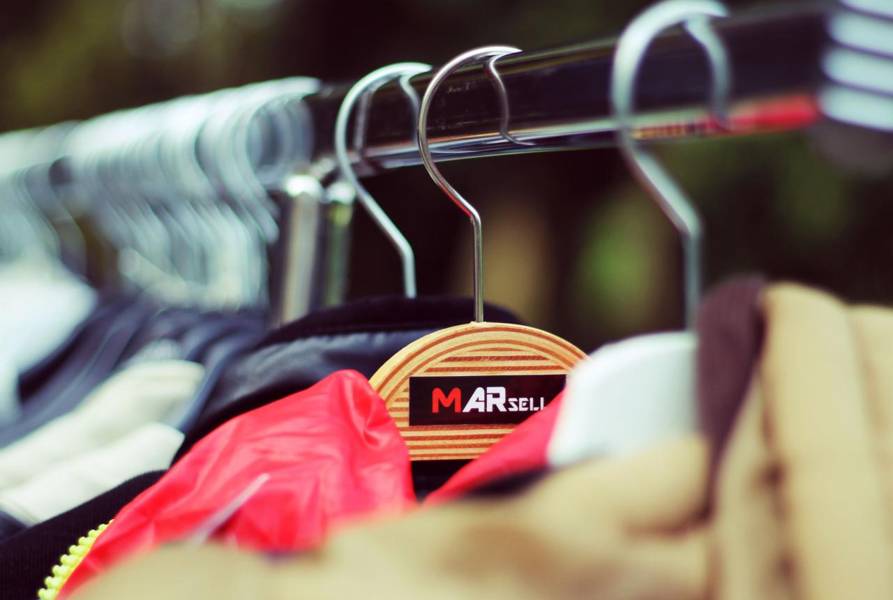 Rząd ubrań wiszących na metalowym wieszaku, z drewnianym wieszakiem z logo 'MARSELI' na pierwszym planie, w naturalnym świetle.