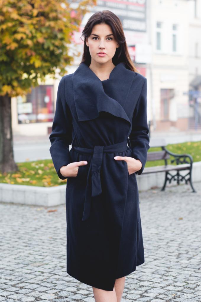 Kobieta w długim, granatowym płaszczu z szerokim kołnierzem i paskiem, pozująca na tle miejskiego parku z ławką i drzewem.