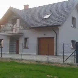 Dwukondygnacyjny dom murowany z szarym tynkiem, ciemną dachówką i balkonem z metalową balustradą, otoczony siatką ogrodzeniową, z garażem z brązową bramą segmentową.