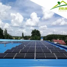 Instalacja fotowoltaiczna na niebieskim dachu przemysłowym, widok z góry na panele słoneczne w regularnych rzędach, w tle zabudowania wiejskie i błękitne niebo z chmurami, w prawym górnym rogu logo...