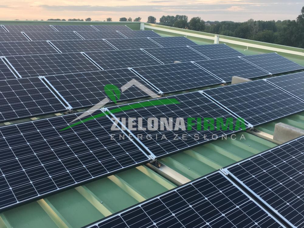 Rząd paneli słonecznych na zielonym dachu z blachy trapezowej, widoczne betonowe podpory i logo firmy na pierwszym planie.