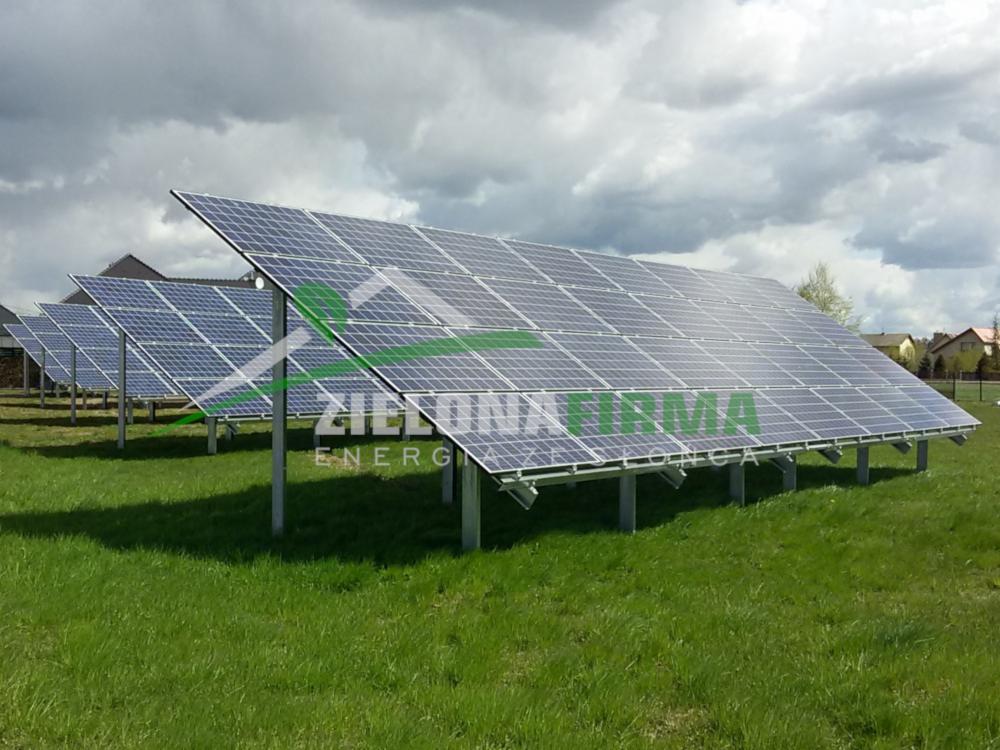Rząd instalacji paneli słonecznych na metalowych konstrukcjach, widoczne budynki mieszkalne w tle, pochmurne niebo.