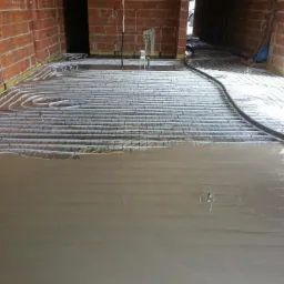 Rozlana wylewka na rurach ogrzewania podłogowego, widoczne fragmenty siatki zbrojeniowej i ceglane ściany w surowym stanie.