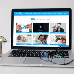 Laptop wyświetlający stronę internetową z różnorodnymi grafikami, okulary na klawiaturze, kubek z kwiatowym wzorem i książka obok, na białym biurku.