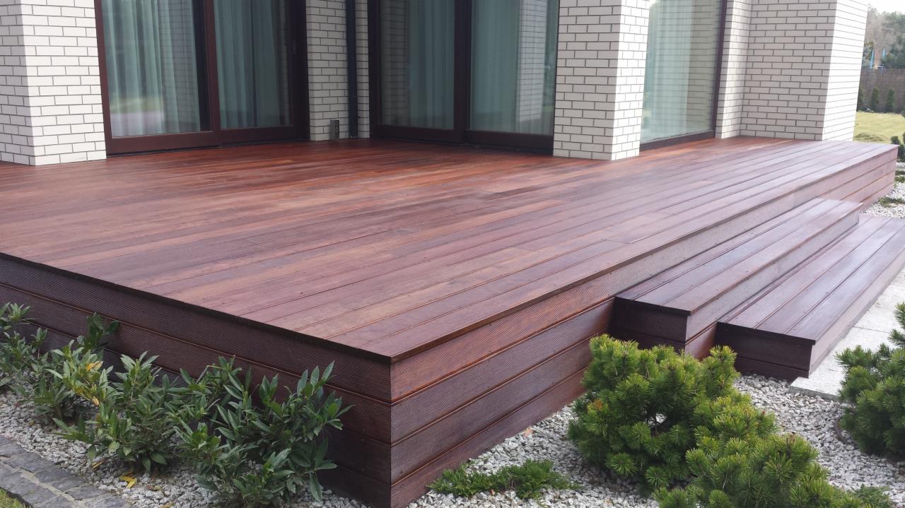 Ciemnobrązowy taras z drewna kompozytowego z dwoma stopniami, otoczony białym kamieniem i zielonymi krzewami, przylegający do jasnego budynku z cegły.