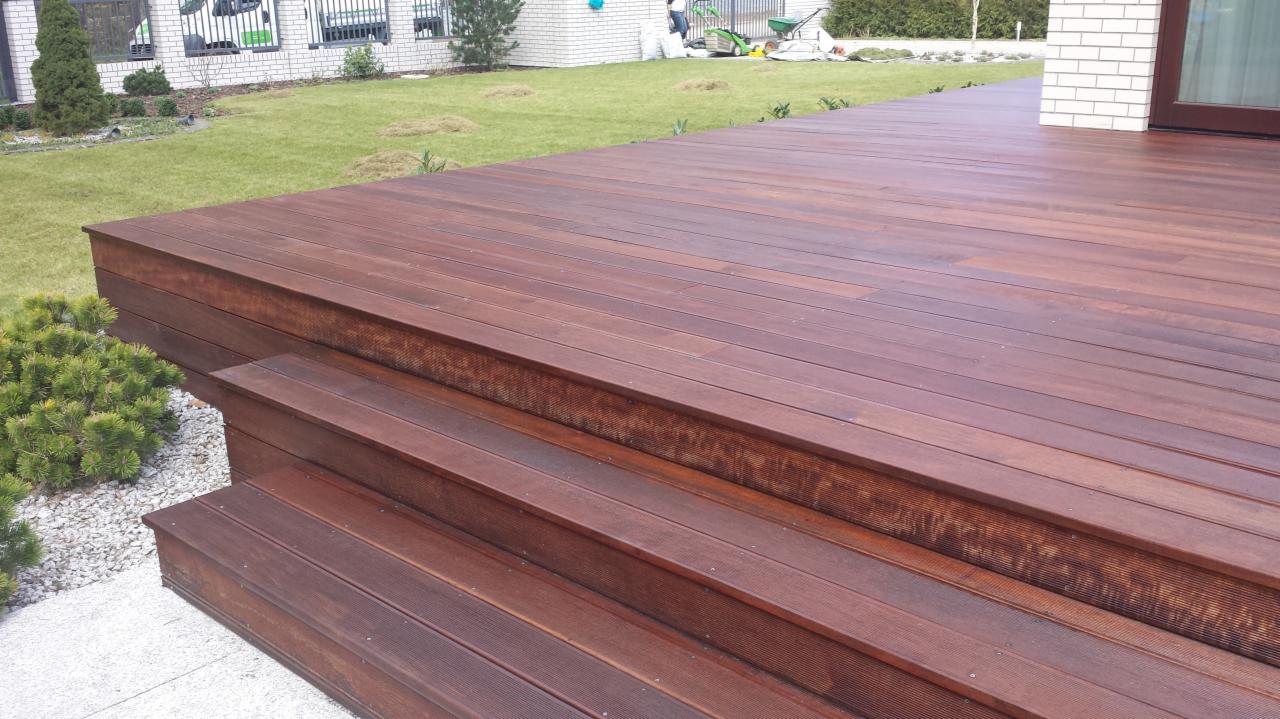 Ciemnobrązowy taras z drewna egzotycznego, z niskimi schodami, prowadzący do drzwi balkonowych w białej cegle, otoczony zielonym trawnikiem i krzewami.