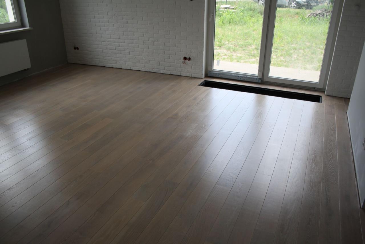 Jasny, drewniany parkiet w nowym pomieszczeniu z białymi ścianami z cegły i dużym oknem balkonowym, widoczny fragment czarnej kratki wentylacyjnej w podłodze.