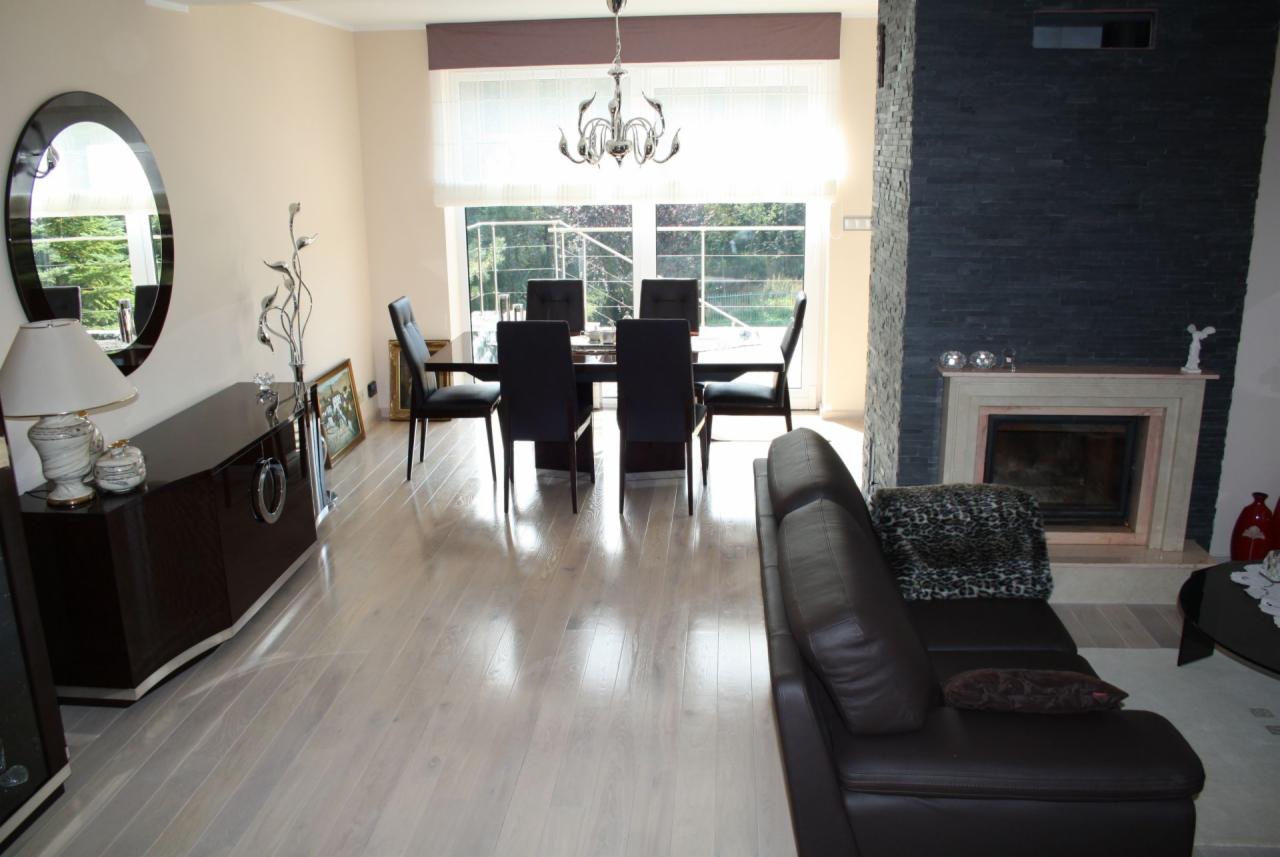 Jasna, lakierowana podłoga z desek w przestronnym salonie z ciemnymi meblami, kominkiem z czarnego kamienia i dużym oknem balkonowym.