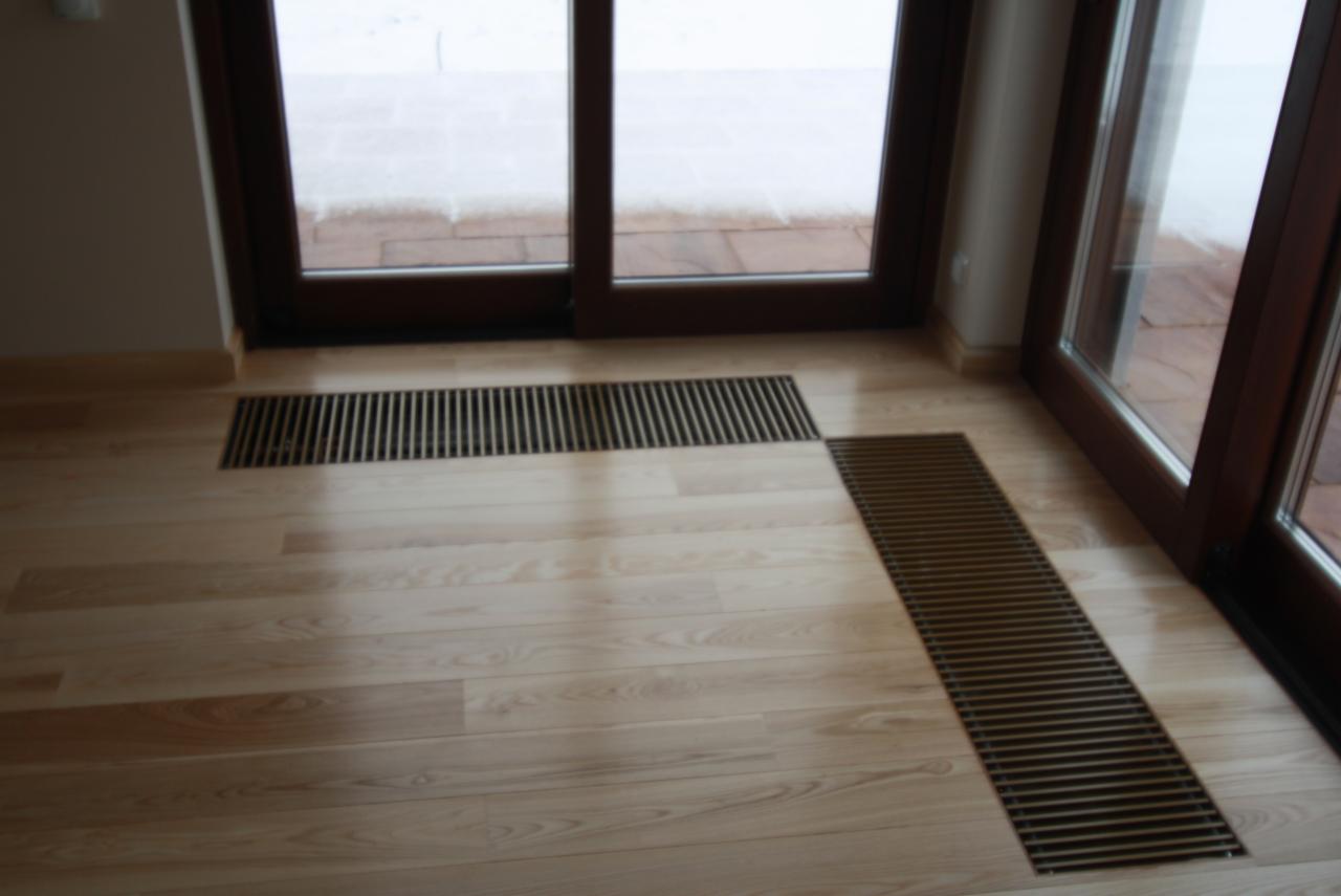 Jasna drewniana podłoga z dwiema kratkami wentylacyjnymi wpuszczonymi w podłogę, widok na taras przez duże okno balkonowe.