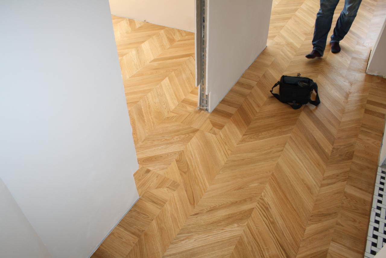 Nowo ułożony parkiet w jodełkę francuską z jasnego drewna, widoczna torba narzędziowa i fragment osoby w jeansach.